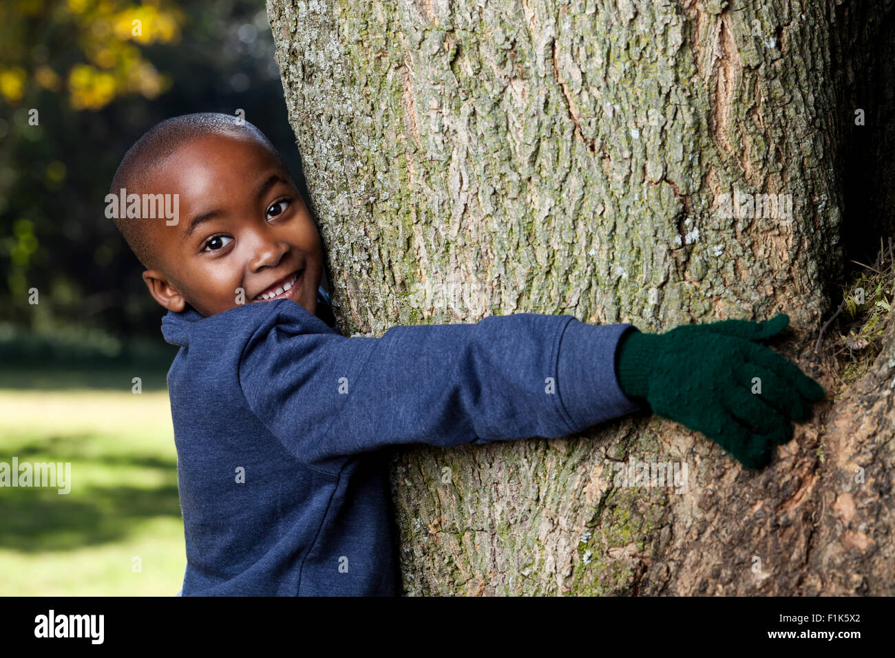 Young African boy hugging un arbre dans un parc tandis que smiling at camera Banque D'Images