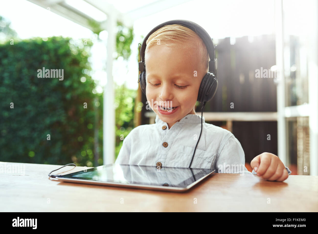 Petit garçon rire heureux d'écouter de la musique sur son tablet pc au moyen d'un casque stéréo comme il est assis à l'extérieur sur une chaude journée d'été Banque D'Images