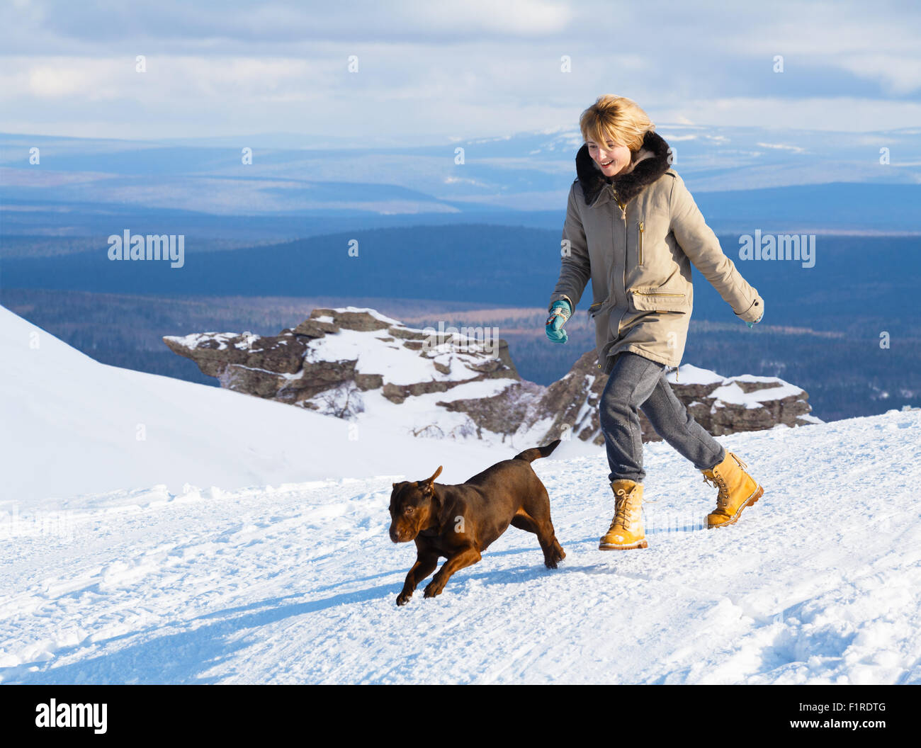 Femme courir et sauter avec chien dans le champ de neige en face de montagnes au loin sur une journée ensoleillée. Jouer, Dog Training, happin Banque D'Images