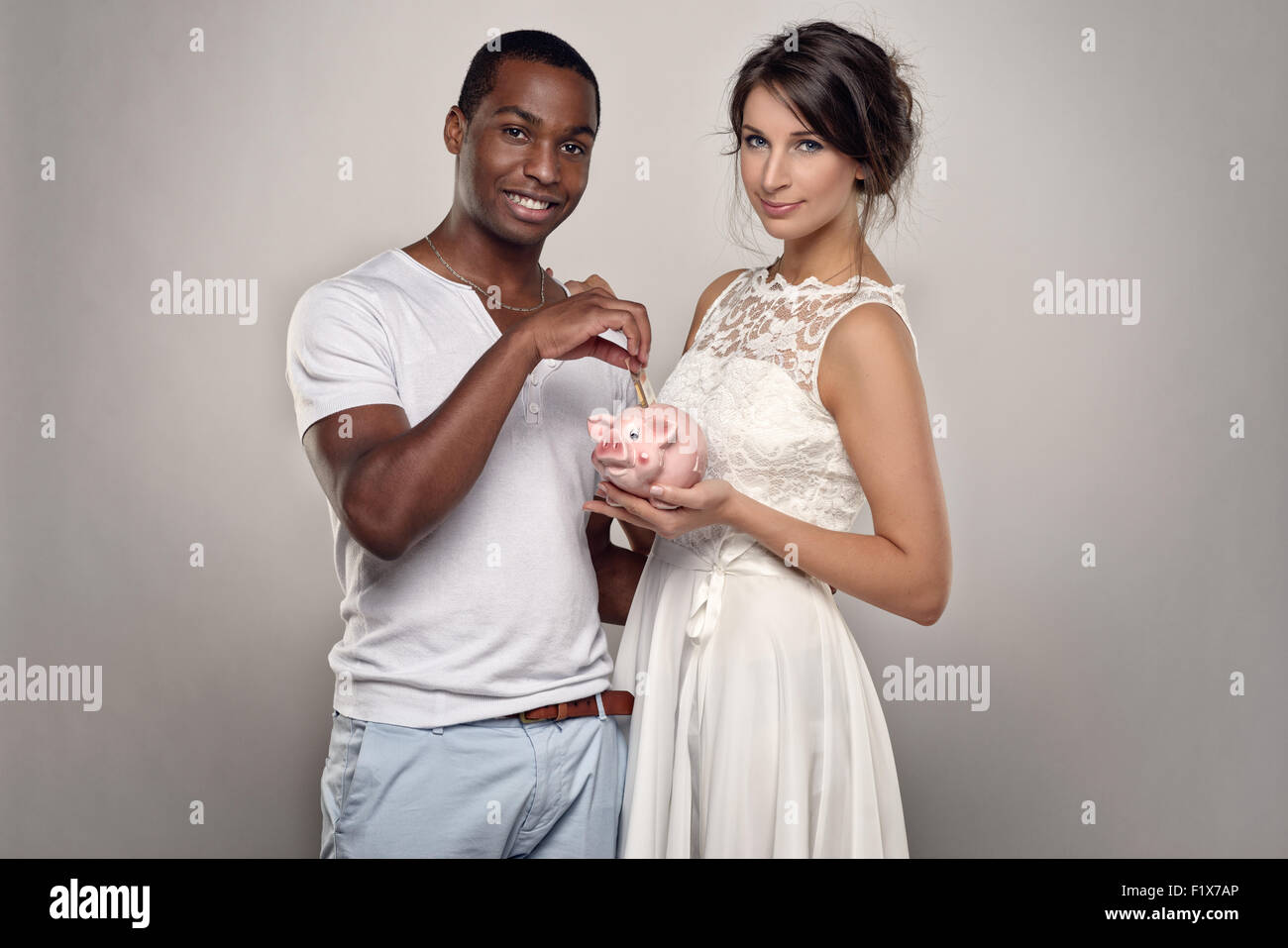 Trois quart d'un jeune couple avec Tirelire souriant à la caméra contre le mur gris. Banque D'Images