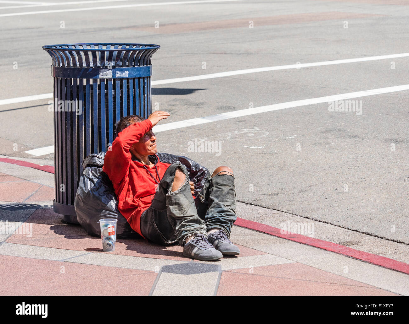 Un sans-abri avec un pantalon déchiré au niveau des genoux est assis sur le trottoir et s'appuie contre une poubelle et retient son bras vers le haut. Banque D'Images