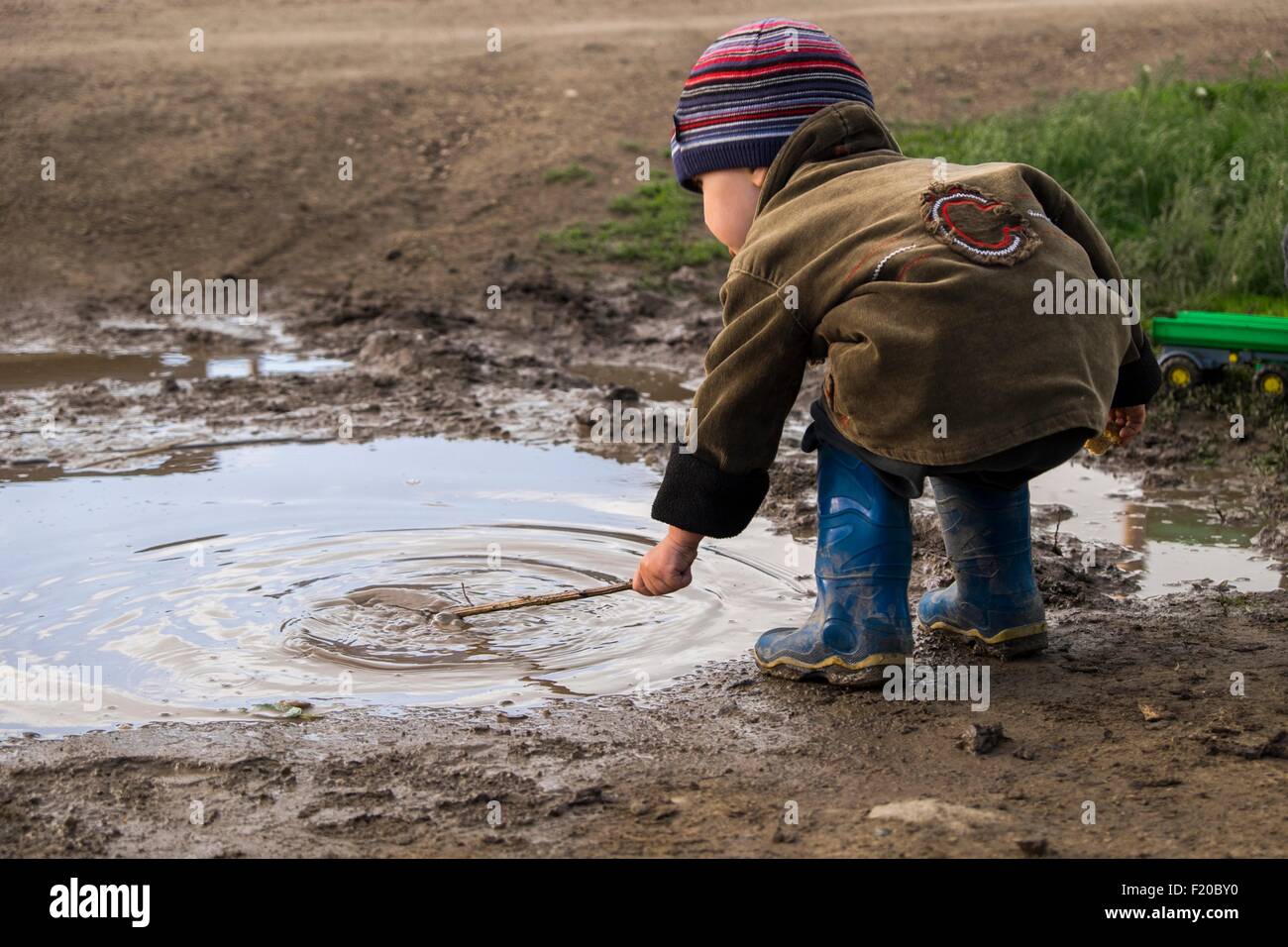 Tout-petit mâle portant des bottes en caoutchouc à jouer avec stick in puddle Banque D'Images
