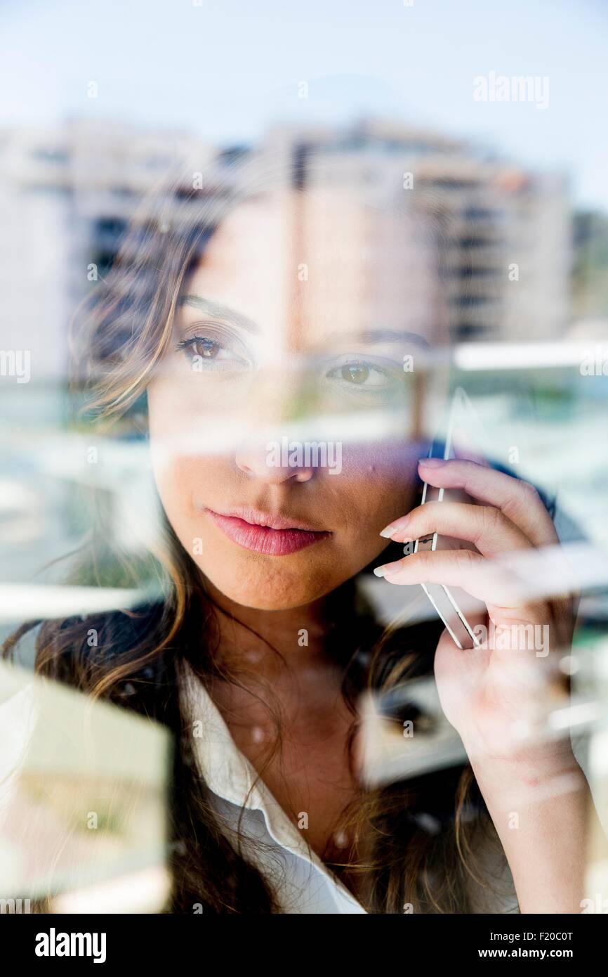 Young woman using smartphone, pensive expression, photographié à travers le verre Banque D'Images