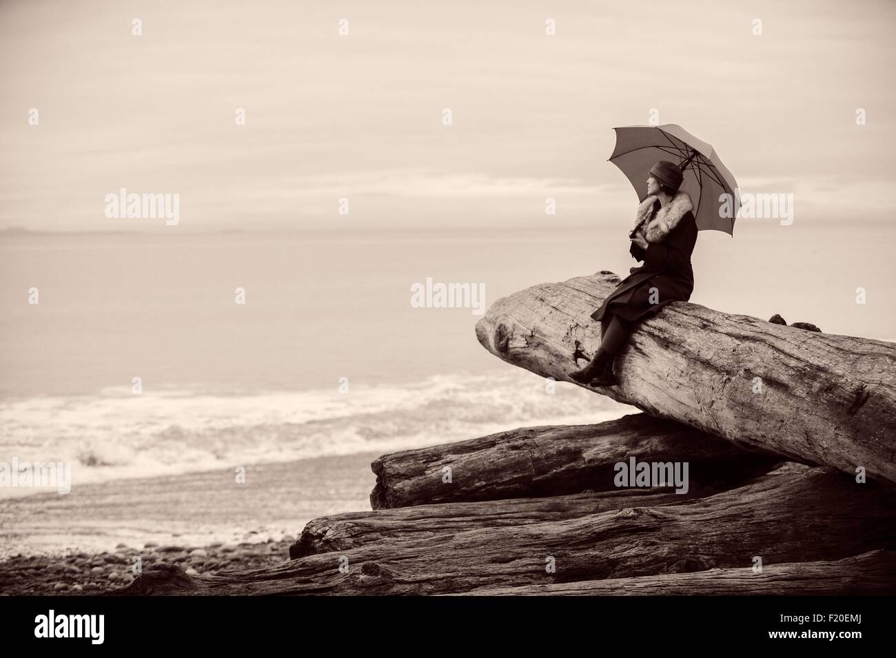 Femme avec parapluie assis sur de gros tronc d'arbre de bois flotté sur la plage Banque D'Images