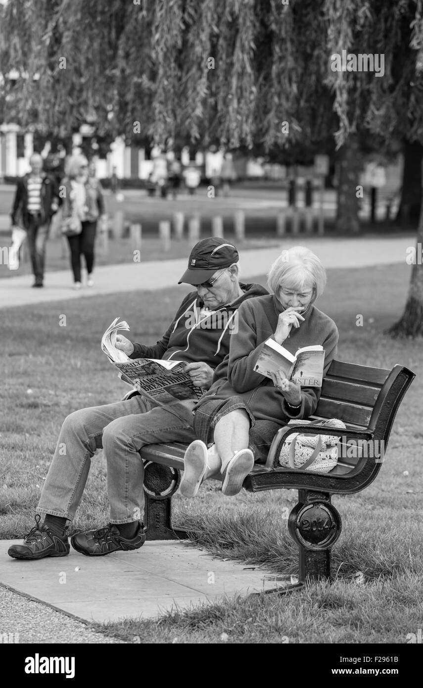 Couple d'âge moyen, la dame de détente avec un livre de poche et l'homme avec un journal sur un siège du parc, England, UK Banque D'Images