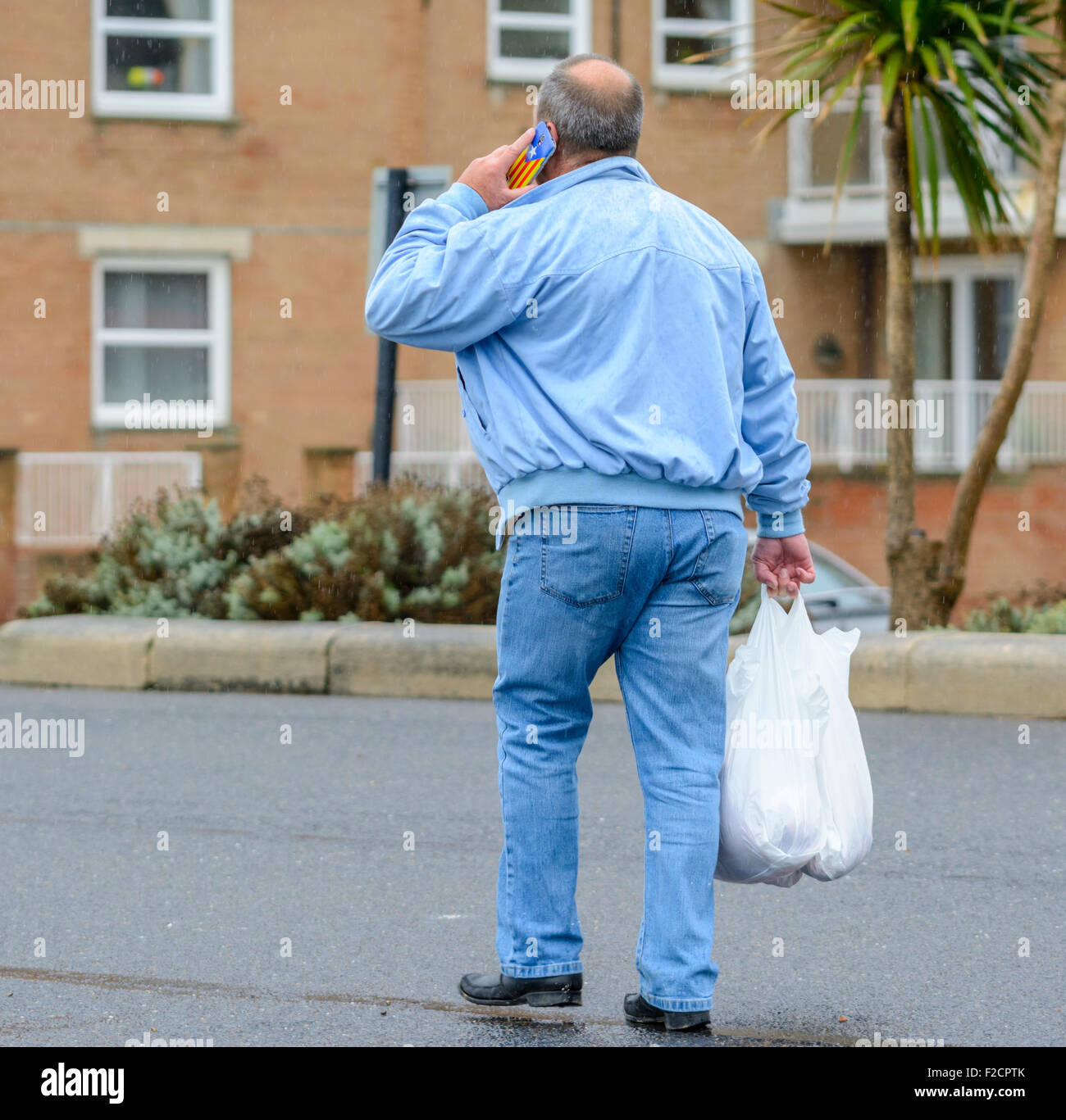 Homme marchant le long d'un chemin transportant des achats et parlant sur un téléphone mobile (téléphone cellulaire ou cellulaire) en Angleterre, au Royaume-Uni. Banque D'Images