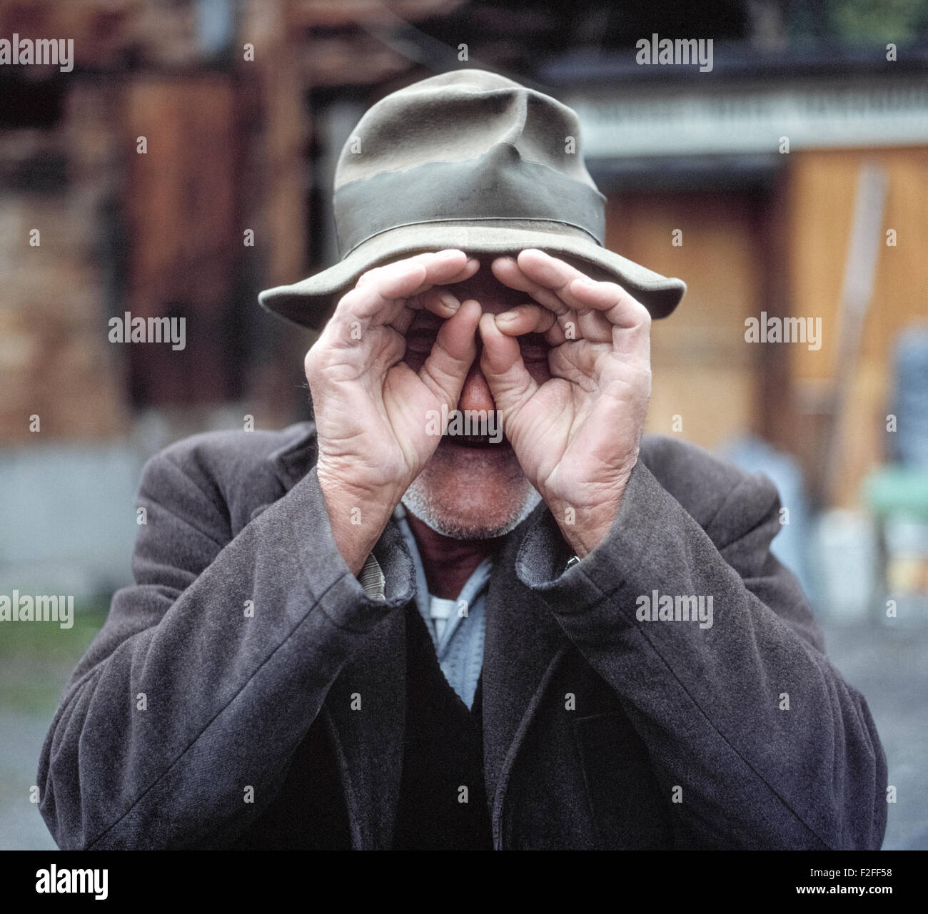 Un vieil homme aux tempes grisonnantes en Suisse café pour la caméra en mettant ses mains à ses yeux pour prétendre qu'il est à la recherche à travers une paire de jumelles pour espionner le photographe. Banque D'Images