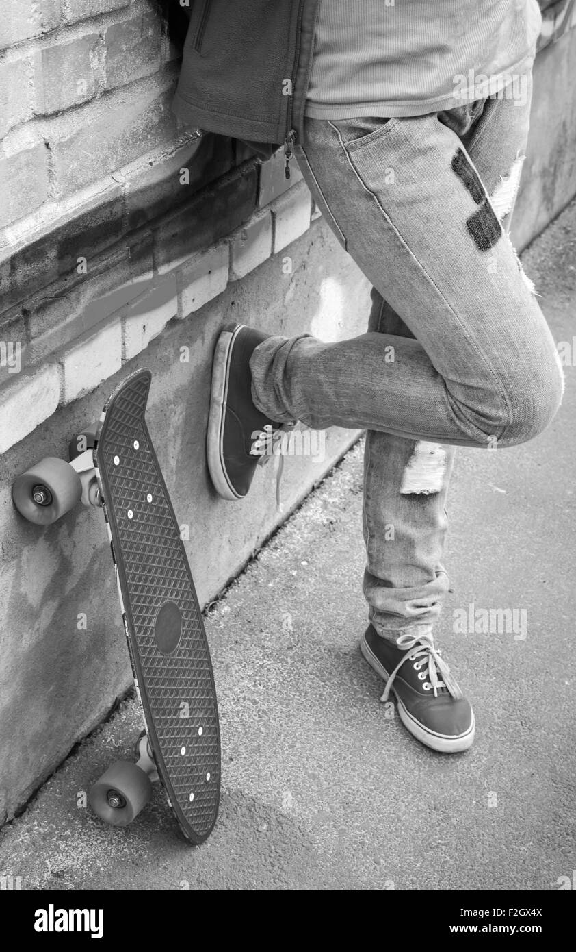 Adolescent en jeans et gumshoes stands avec mur de brique près de skateboard Banque D'Images