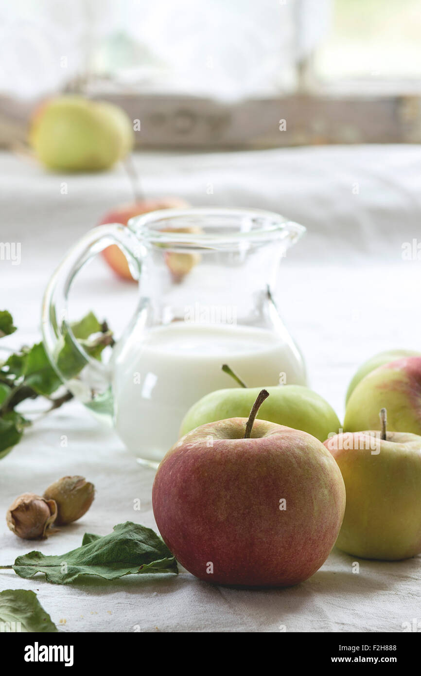 Tas de pommes avec des feuilles, de noisettes et de pot de lait sur nappe blanche avec fenêtre en arrière-plan Banque D'Images