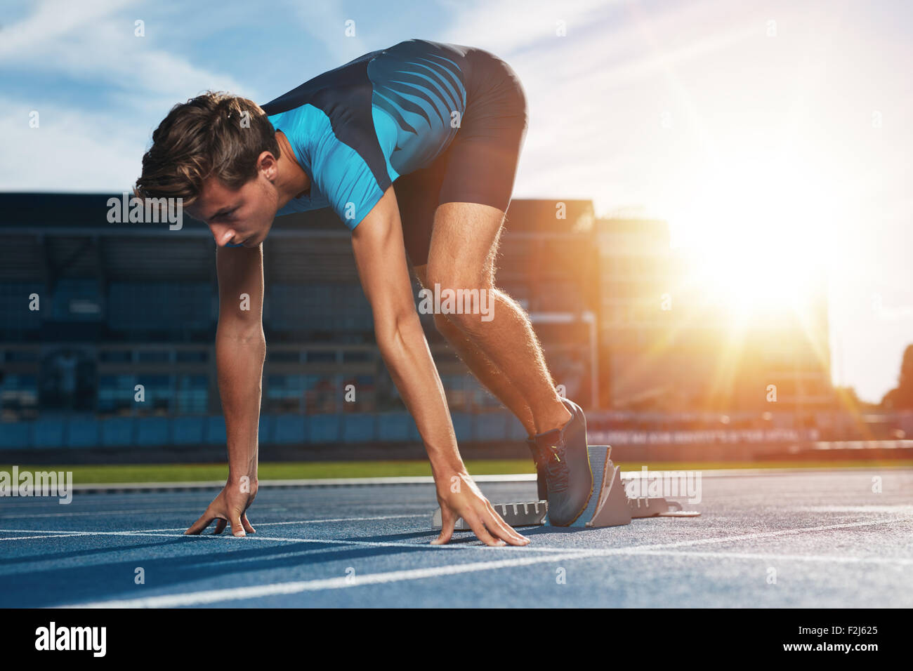 Young male runner en tenant prêt à démarrer position contre la lumière du soleil. Sprinter sur bloc de départ d'une piste d'athlétisme Banque D'Images