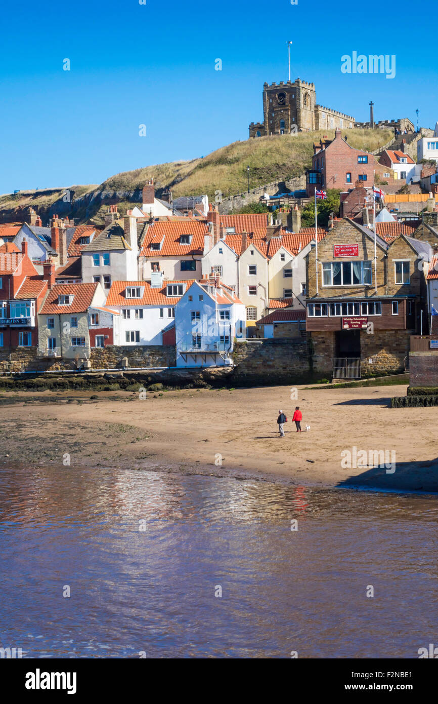 Couple avec un chien sur la plage de Tate à Whitby Harbour avec des chalets sur le côté est de la ville et l'église St Mary à l'automne Banque D'Images