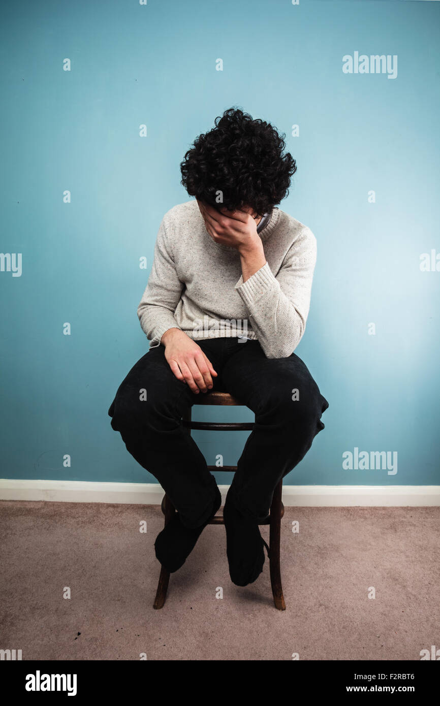 Un jeune homme triste est assis sur une chaise haute par un mur bleu Banque D'Images