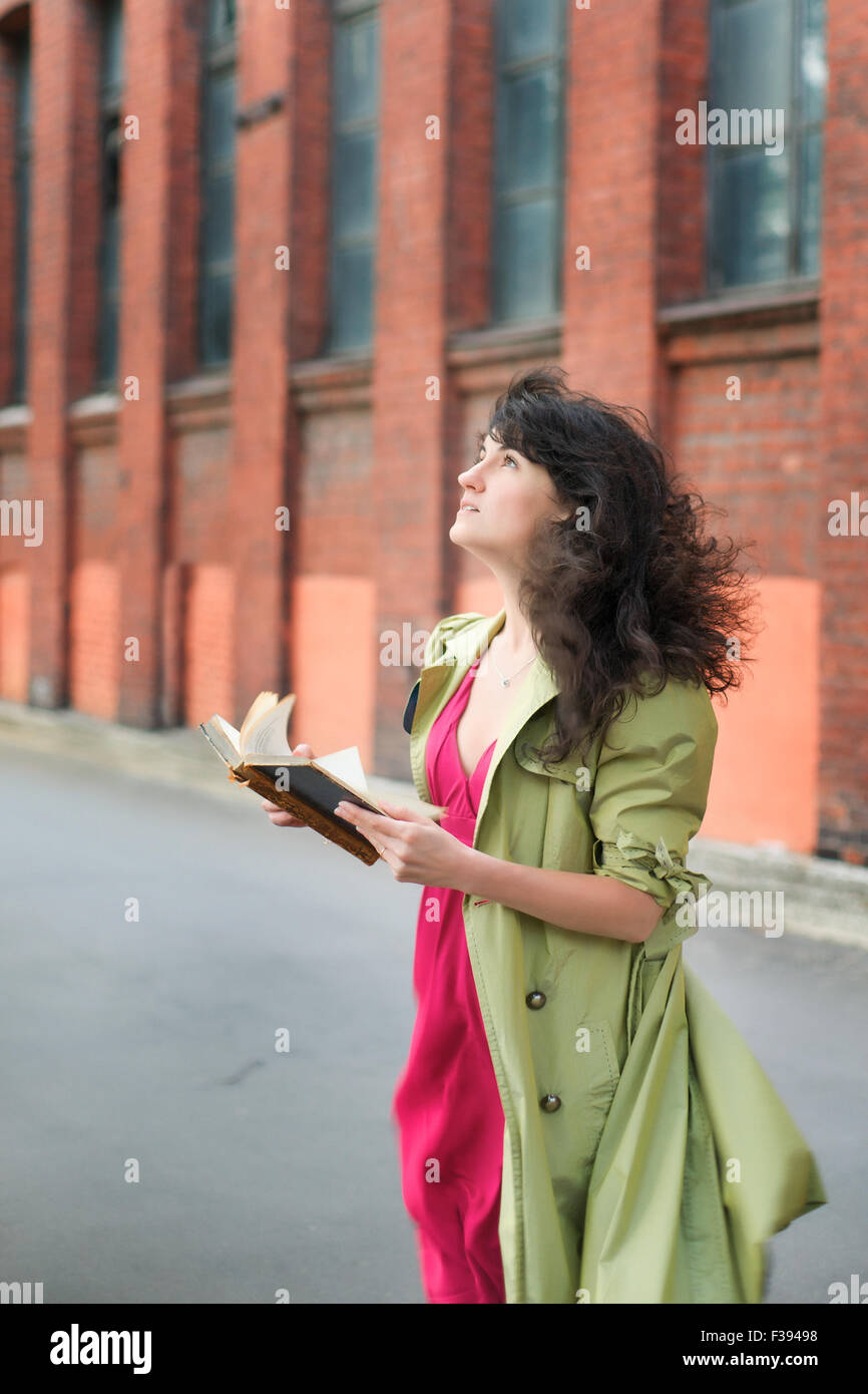 Belle jeune femme avec cheveux voletant dans le vent dans un manteau vert avec de vieux livre sur fond de vintage industrial landsca Banque D'Images