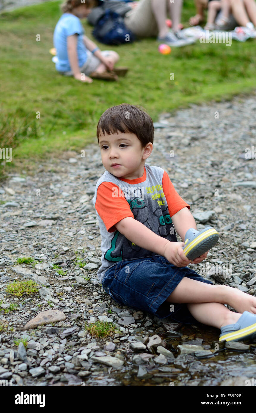 Un jeune garçon est assis sur le sol au bord d'un ruisseau d'obtenir une pierre de sa chaussure. Banque D'Images
