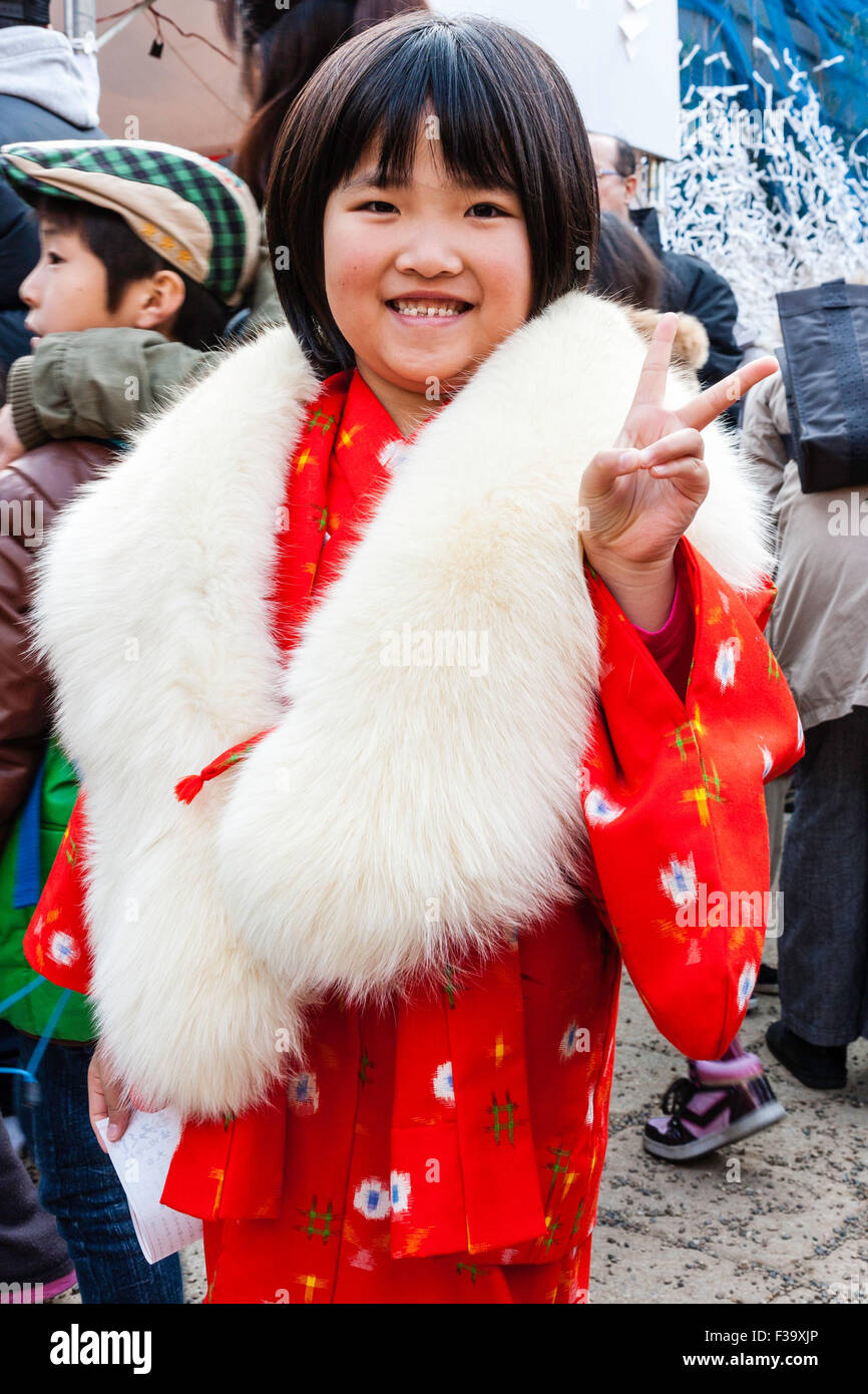 L'enfant japonais, fille, 6-7 ans, de sourire et de donner la paix à des gestes viewer. Porter un kimono rouge avec col de fourrure blanche, traditionnelle au festival times. Banque D'Images