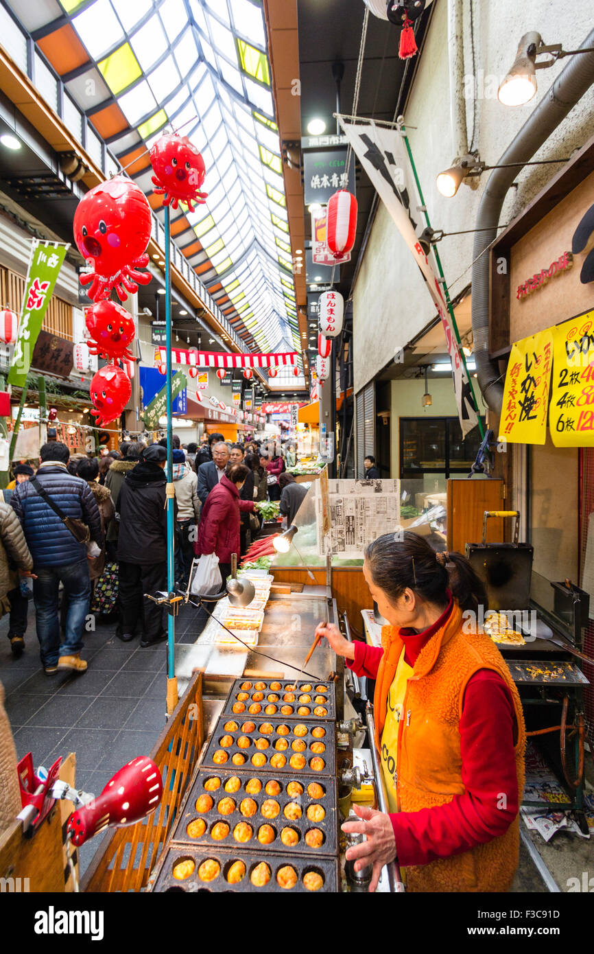 Femme debout à la friture et de la vente des boulettes de poulpe, takoyaki, dans le marché de Kuromon Ichiba à Osaka. Voir l'arcade avec en arrière-plan. Banque D'Images