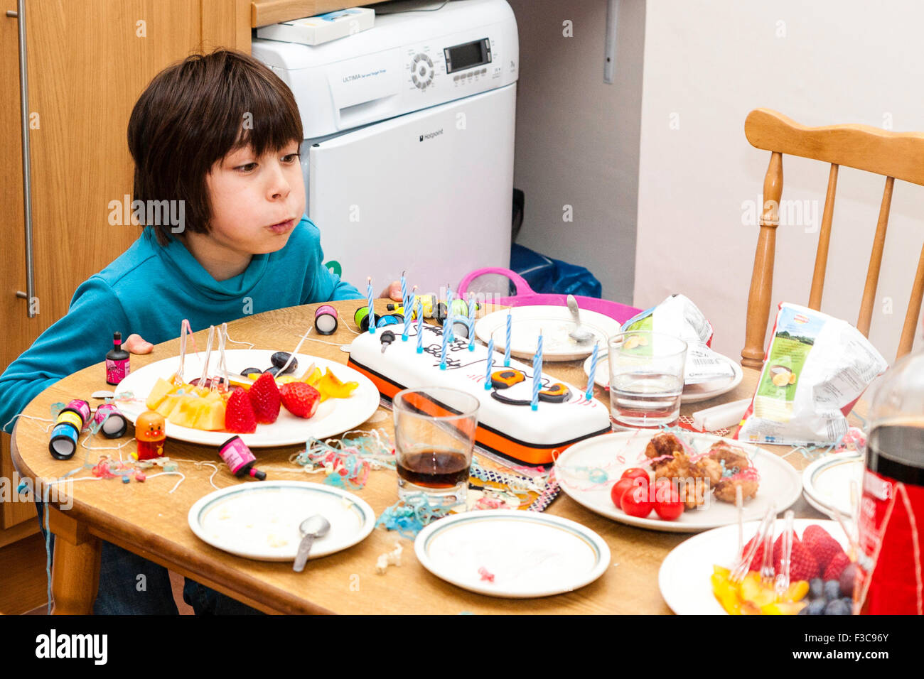 L'origine ethnique mixte (Japonais, Anglais) garçon de 11 ans assis en tête de table de cuisine soufflé les bougies sur le gâteau d'anniversaire. D'autres aliments et boissons. Banque D'Images