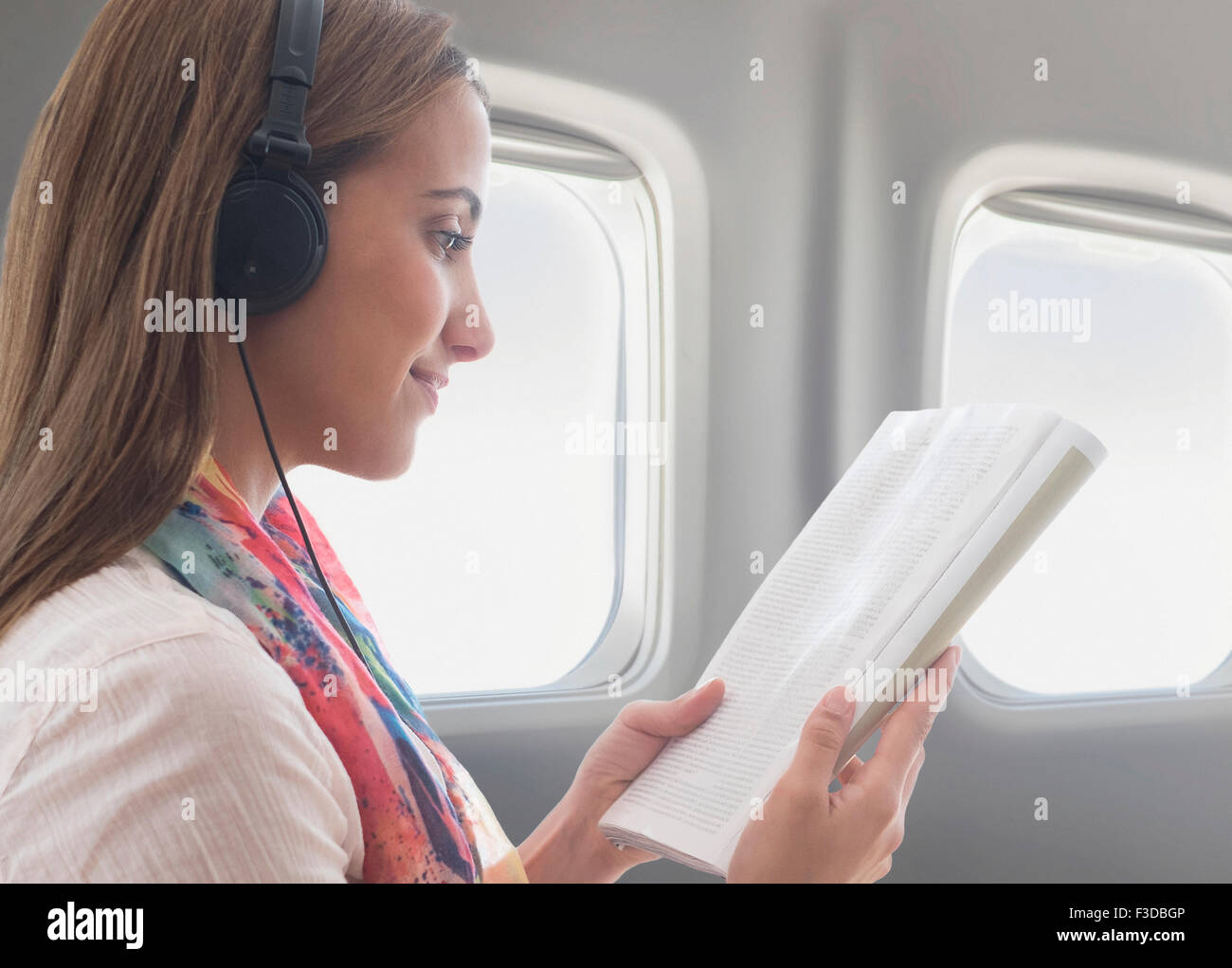 Jeune femme portant des écouteurs lors de la lecture de livre sur l'avion Banque D'Images