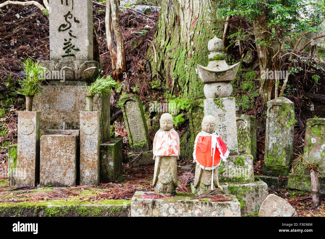Le Japon, Koyasan, Okunoin cemetery. Deux statues jizo bosatsu bibbed rouge placé sur le bloc de pierre avec des pierres tombales et des cèdres arbres derrière. Banque D'Images