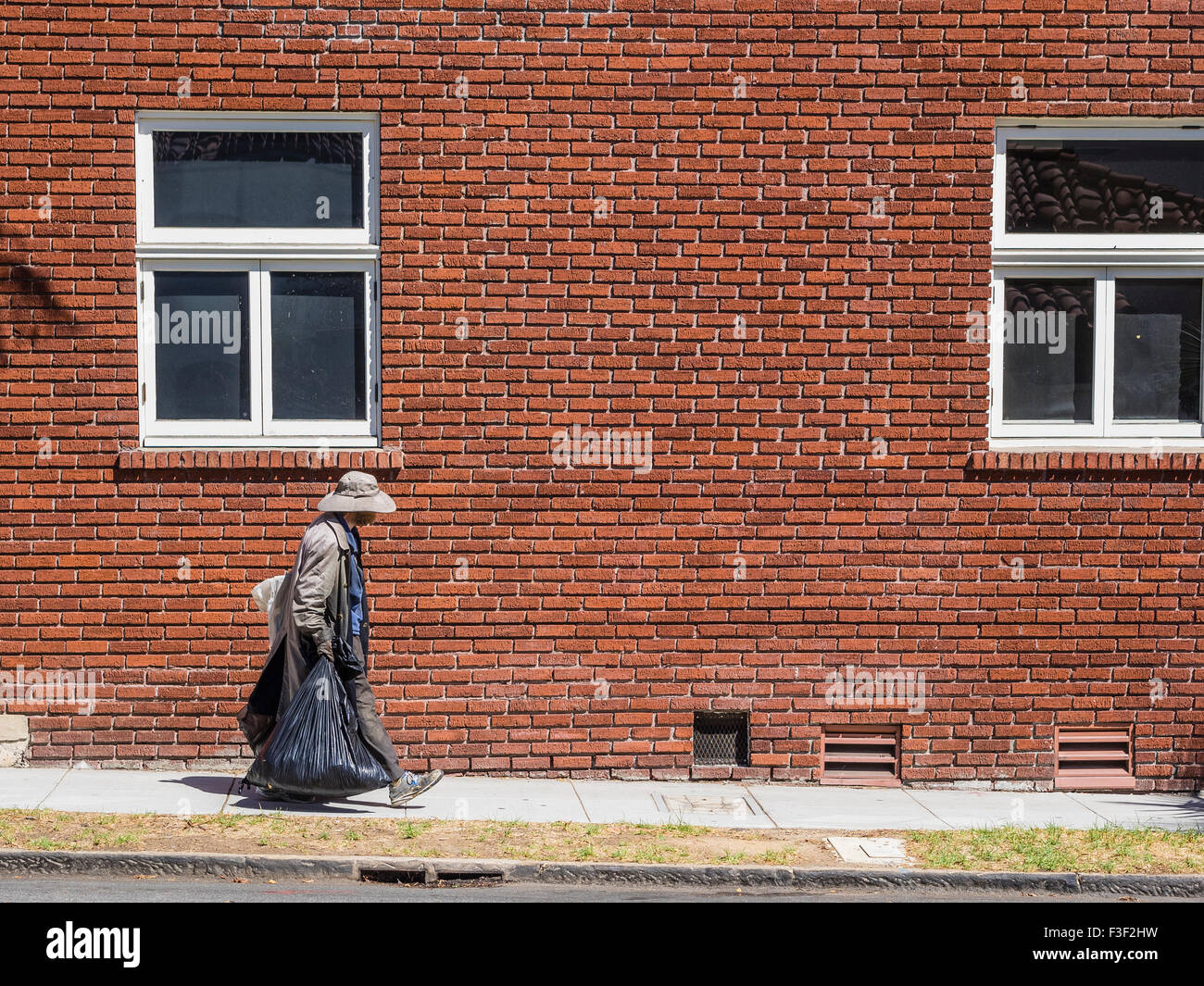Une ancienne sans-abri adultes homme portant un long manteau et un chapeau sur une journée chaude et portant un grand sac poubelle en plastique noir dans chaque ha Banque D'Images