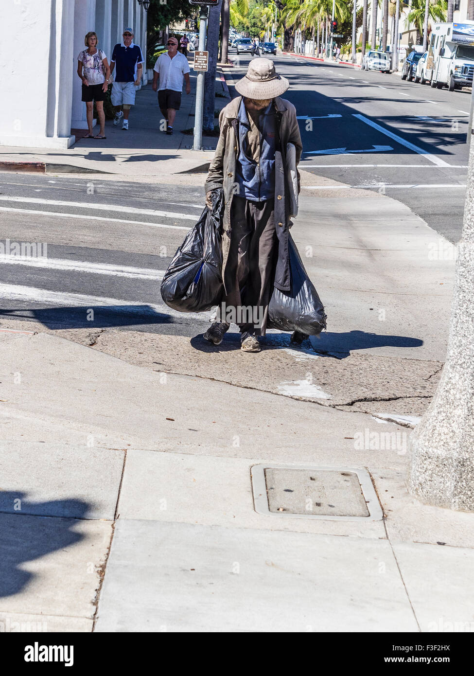 Une ancienne sans-abri adultes homme portant un long manteau et un chapeau sur une journée chaude et transportant deux grands sacs à ordures en plastique noir. Banque D'Images