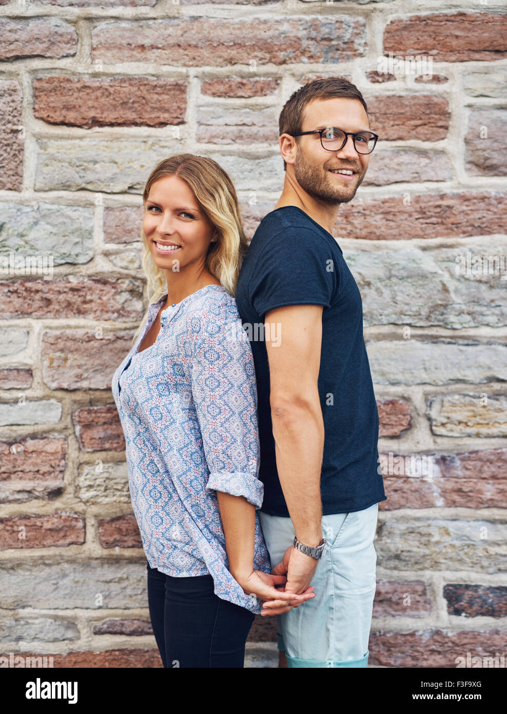 Trois quart Shot of a Happy Young Couple dos à dos tout en se tenant la main et souriant à la caméra contre Bri Banque D'Images