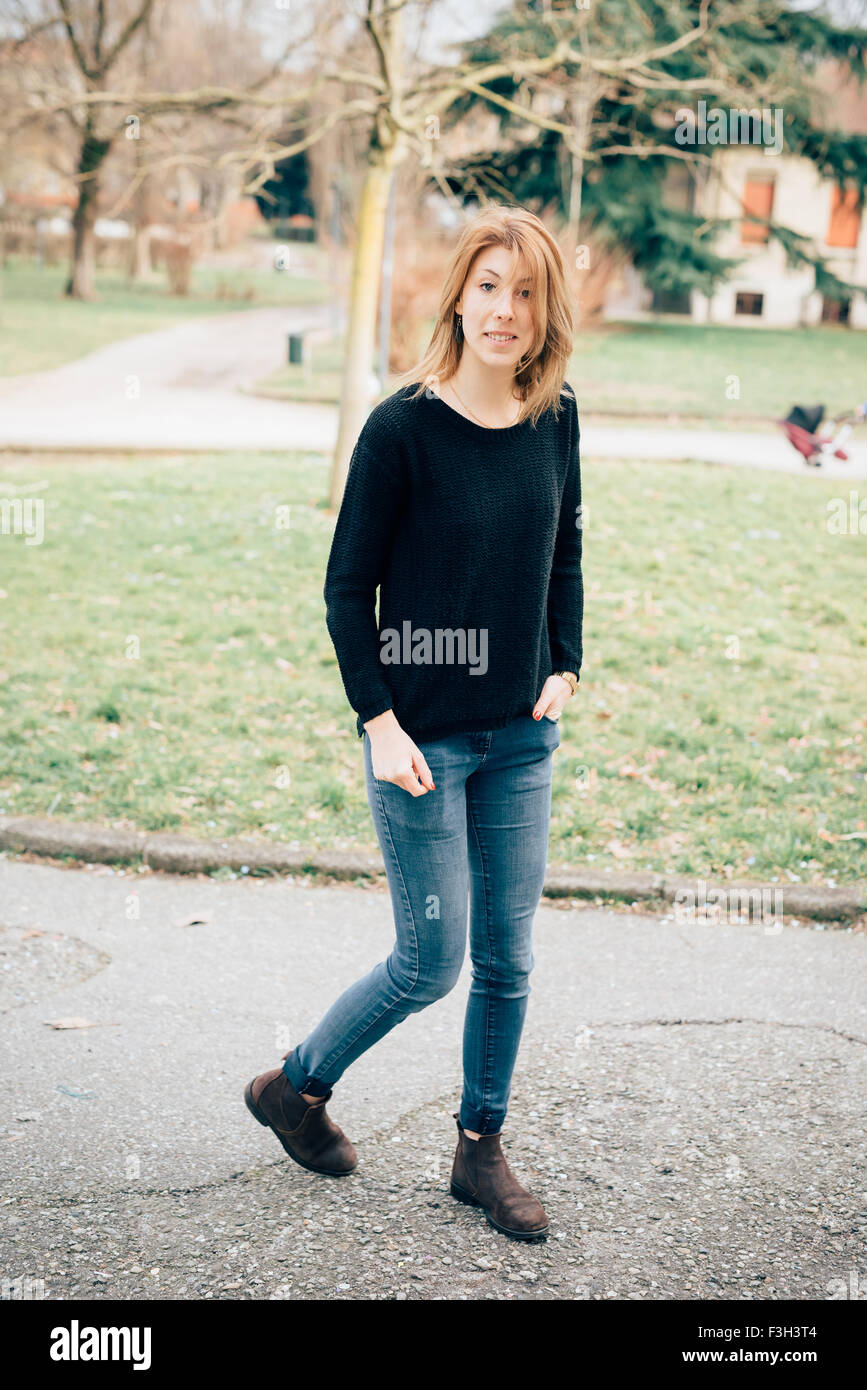 Jeune belle blonde sportive hipster femme marche dans un parc de la ville,  main dans la poche, porter un jean bleu et pull noir, à la caméra en  souriant, - insouciance, les