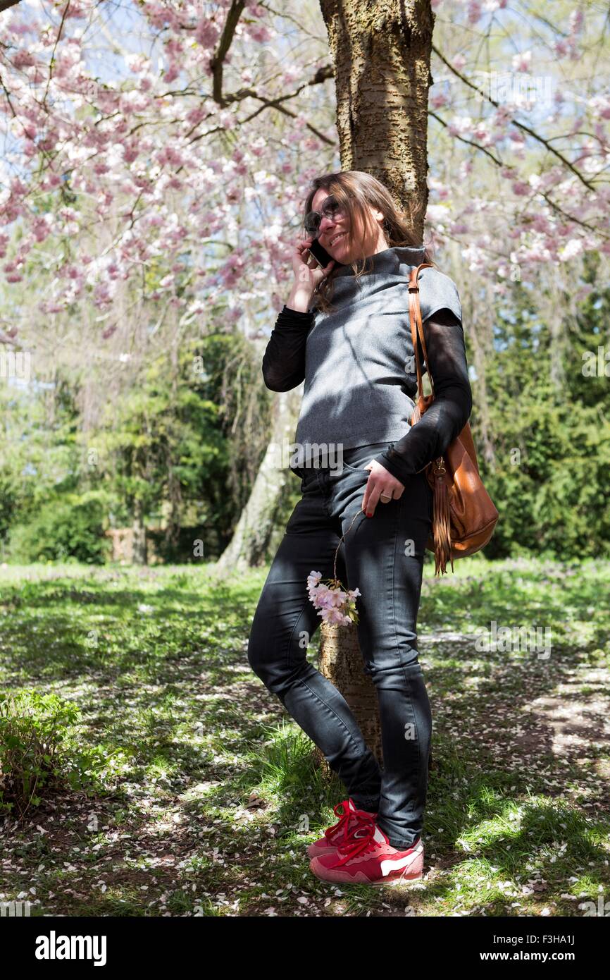 Young woman chatting on smartphone tout en s'appuyant contre l'arbre du parc Banque D'Images