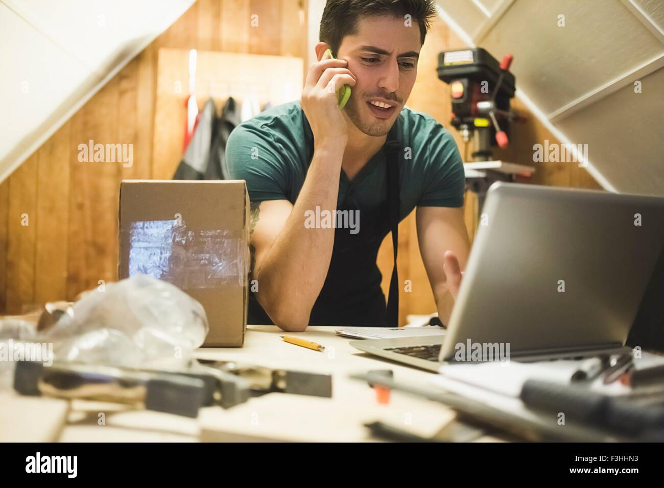 Jeune homme en atelier sitting at desk talking on telephone looking at laptop Banque D'Images