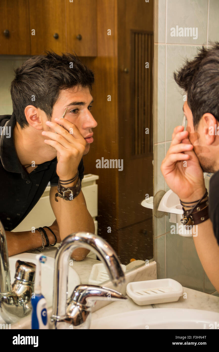 Close Up of Attractive jeune homme aux cheveux noirs lui-même - toilettage à plumer des sourcils avec une pince à épiler les poils errants tout en regardant dans Banque D'Images