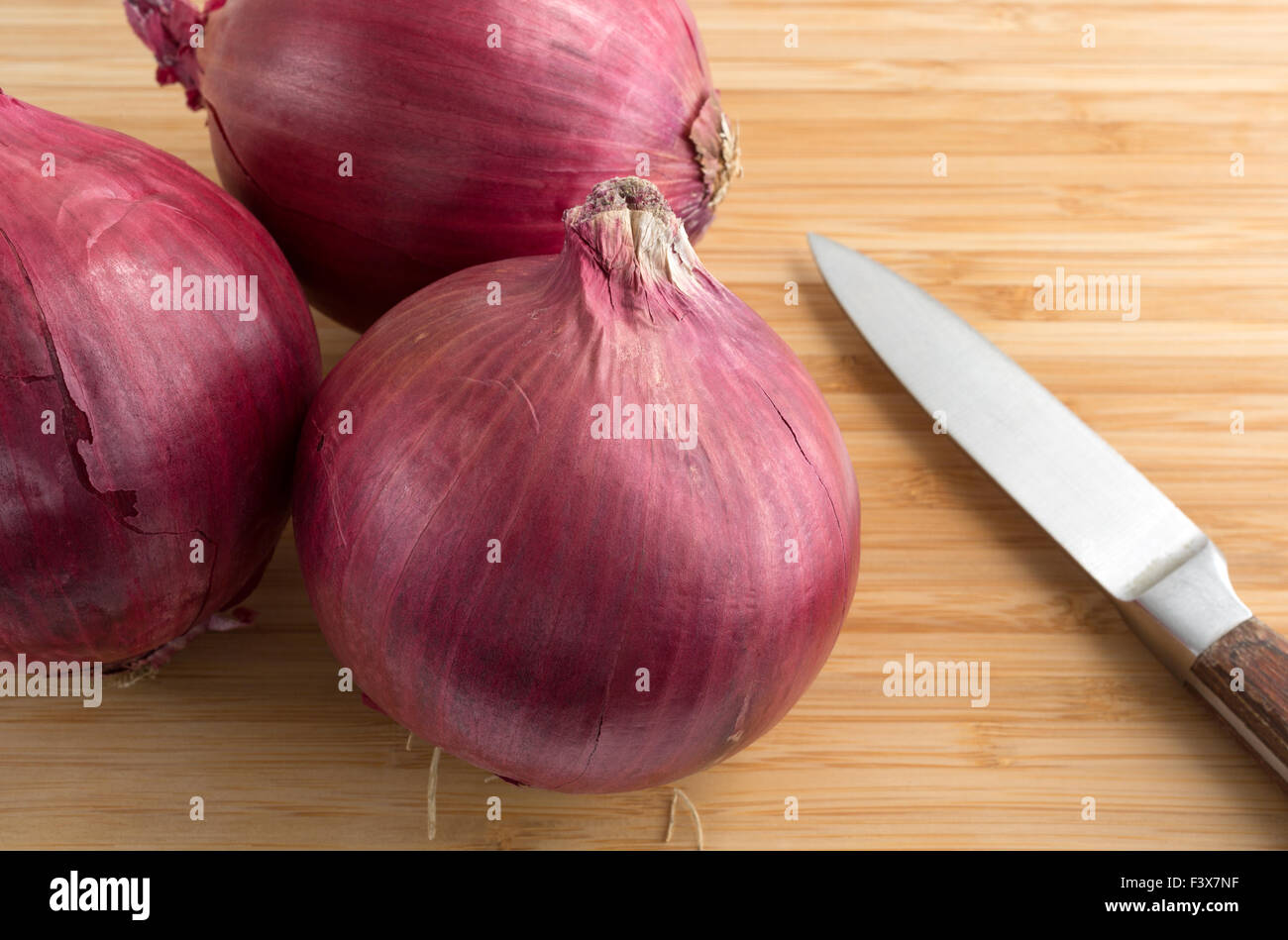 Gros oignons rouges sur une planche à découper en bois avec un couteau sur le côté éclairé par la lumière naturelle. Banque D'Images