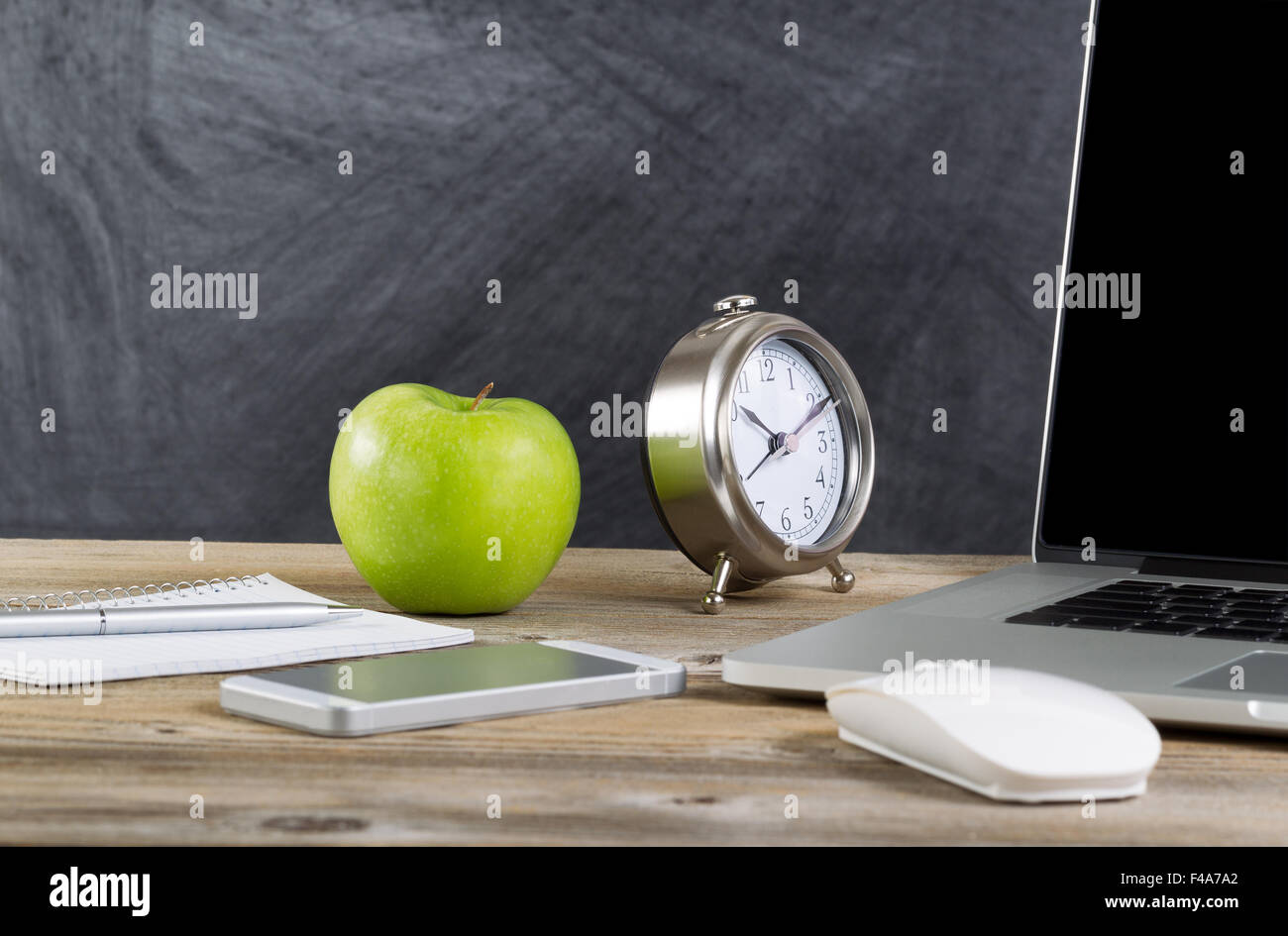 Bureau de l'école avec ordinateur portable, souris, réveil, téléphone cellulaire, ordinateur portable, plume et pomme verte en face du tableau noir. La mise en page à l'horizontale Banque D'Images