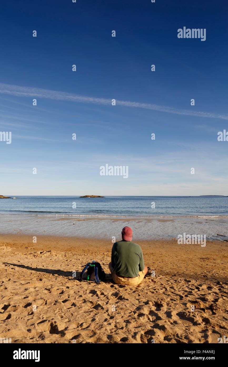 Homme portant des shorts baseball cap à manches longues assis sur une plage de sable fin face à la mer l'Acadia National Park Maine copyspace Banque D'Images