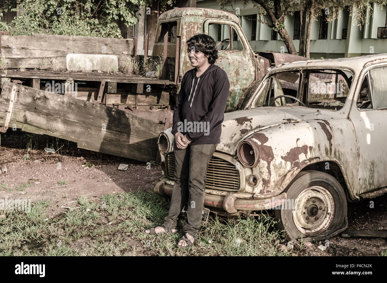 Modèle masculin pose appuyée sur le capot d'une vieille voiture rouillée en ferraille. Banque D'Images