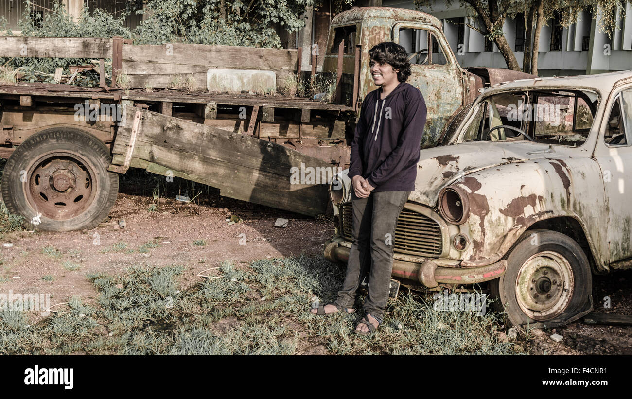 Boy posing in front, appuyé sur le capot d'une vieille voiture rouillée en casse. Banque D'Images