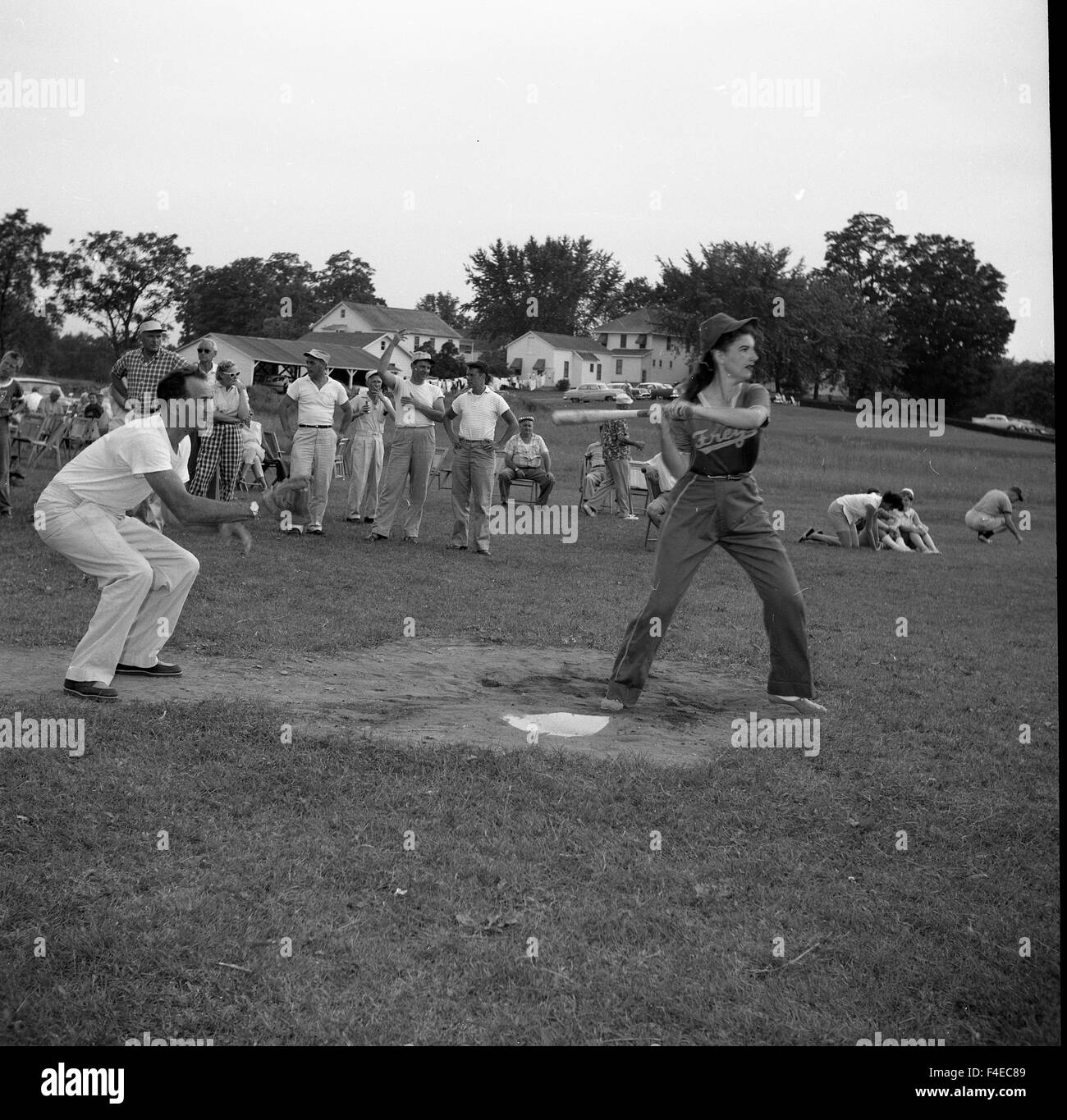 Femme avec batte de baseball à la batte dans un champ dans l'été avec l'homme jouant catcher et d'autres observateurs habillé en 1950 fashions Banque D'Images