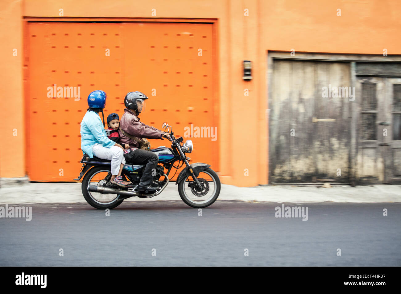 Asian family riding avec enfant looking at camera. Trois personnes sur une moto avec un mur orange règles asiatique mode de transport Banque D'Images