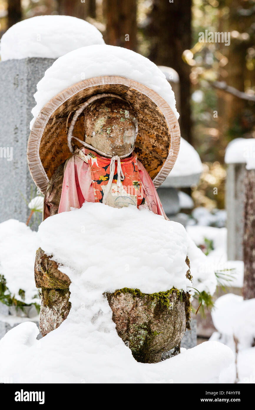 Le Japon, Koyasan, Okunoin cemetery. Assis en pierre couverte de neige-Jizo bosatsu statue bouddhiste, rouge et chapeau de paille conique de soumission. L'hiver Banque D'Images