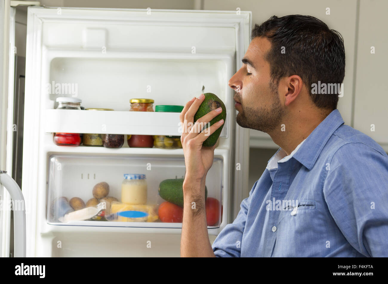 Hispanic male wearing blue shirt debout dans un réfrigérateur odeur d'ouverture un avocat et fermant les yeux Banque D'Images