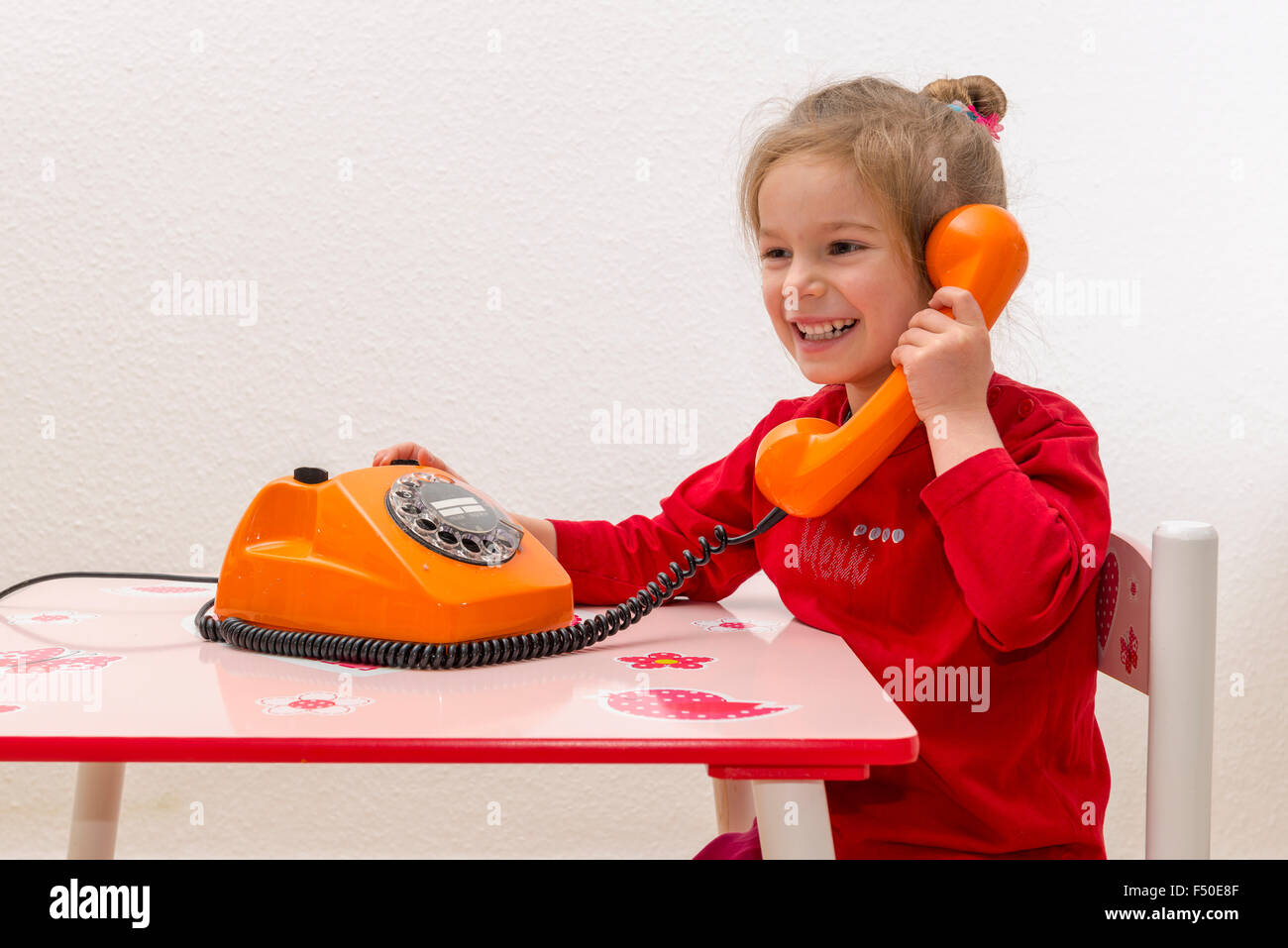 Une blonde fillette de trois ans est assis à une table et l'utilisation d'un téléphone orange, smiling Banque D'Images