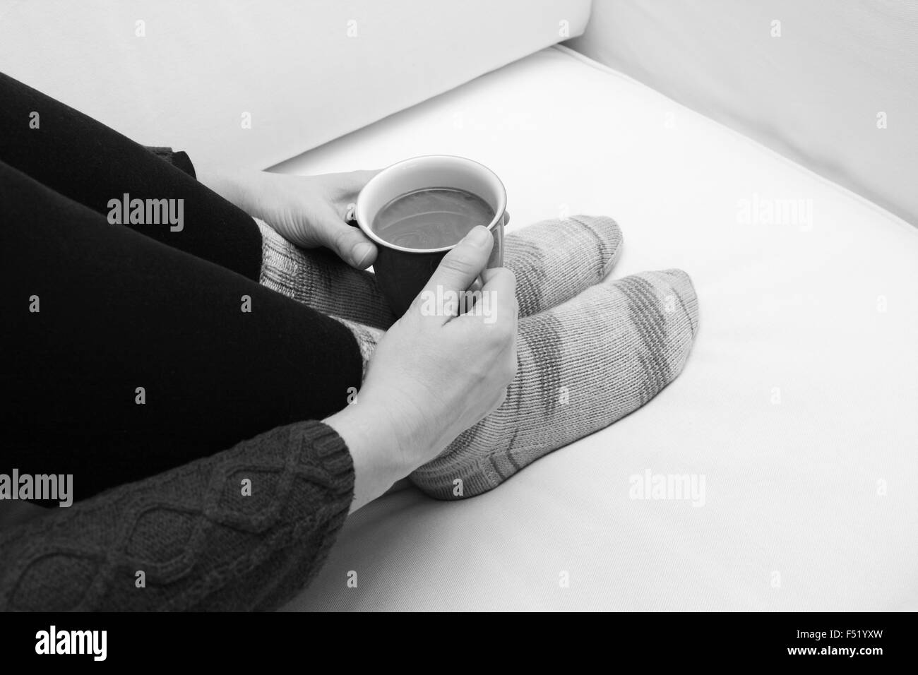 Woman's hands holding une tasse de café chaud ou un thé, recroquevillée sur le canapé, portant des chaussettes tricotées à la main confortable - traitement monochrome Banque D'Images