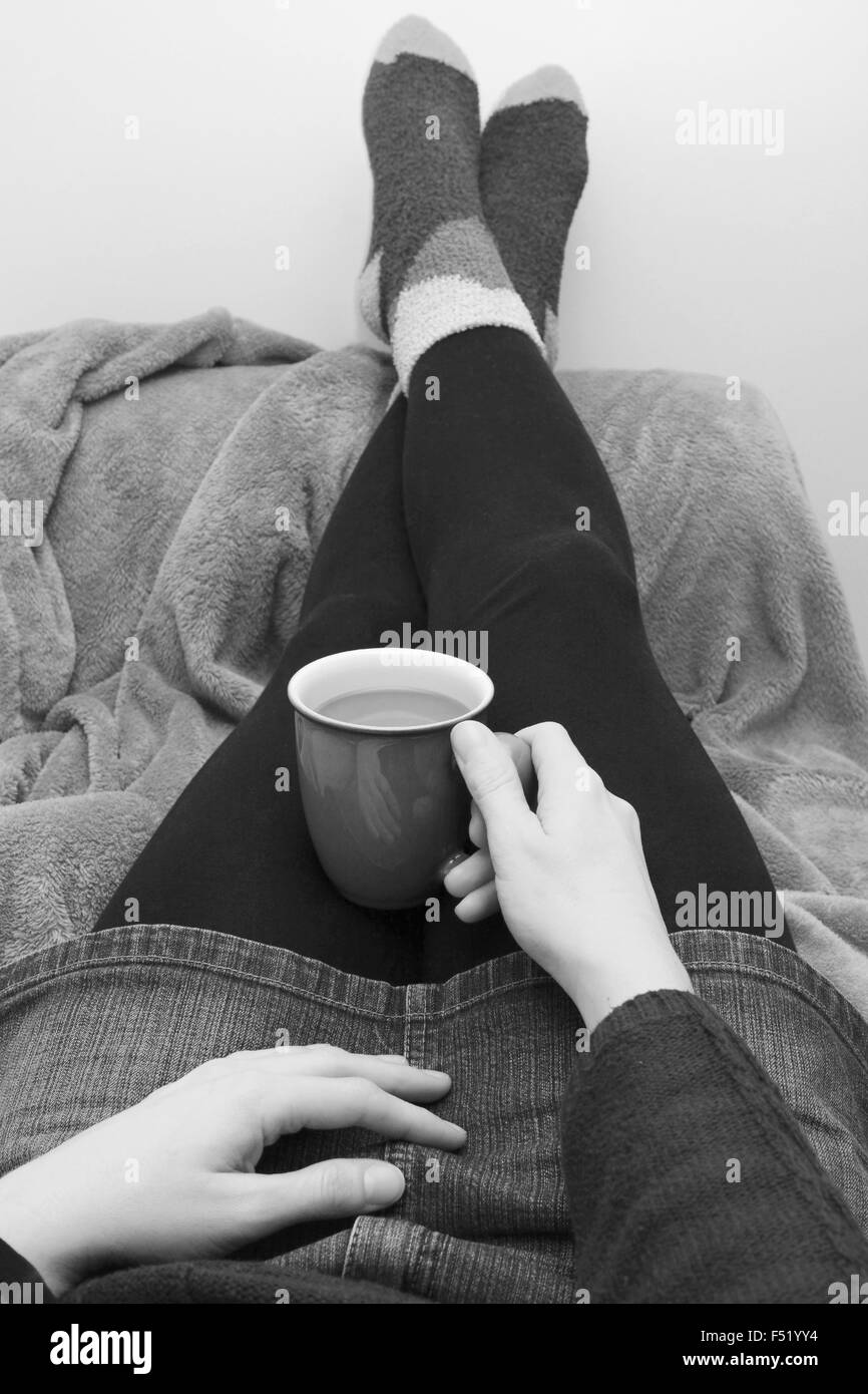 Femme vêtue chaudement, tenant un café chaud ou un thé, avec ses pieds sur le canapé - traitement monochrome Banque D'Images