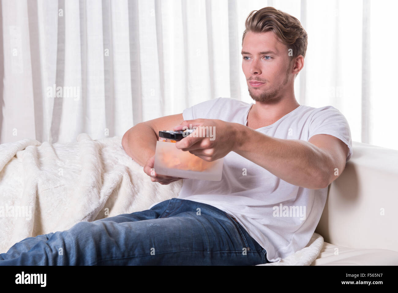 Portrait young man sitting on couch et eating chips et télévision zapping Banque D'Images