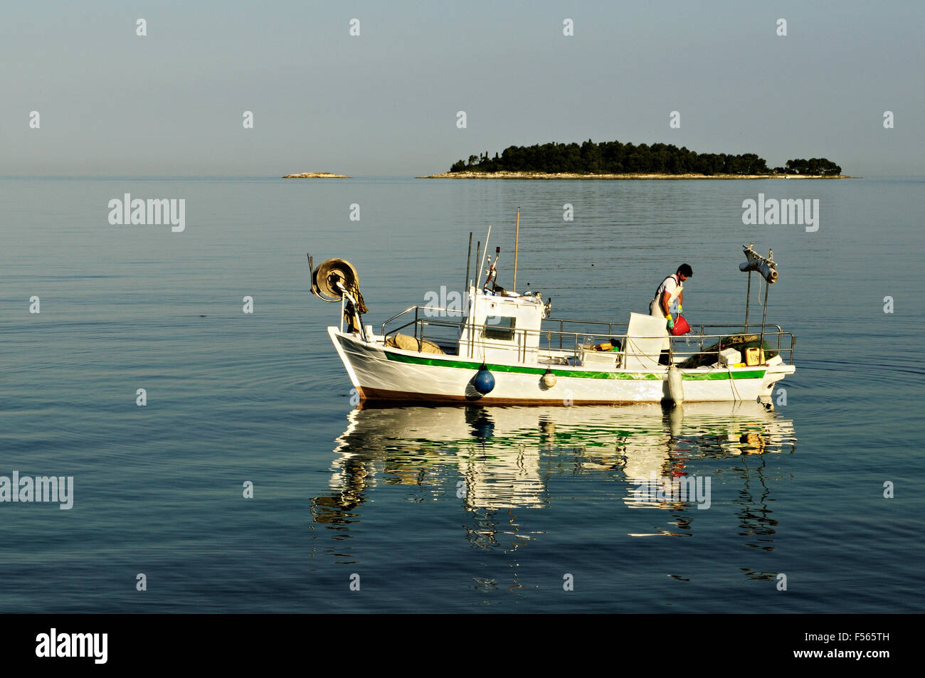 Pêcheur sur son bateau blanc avec une petite île dans l'arrière-plan au lever du soleil, Rovinj, Istrie, Croatie Banque D'Images