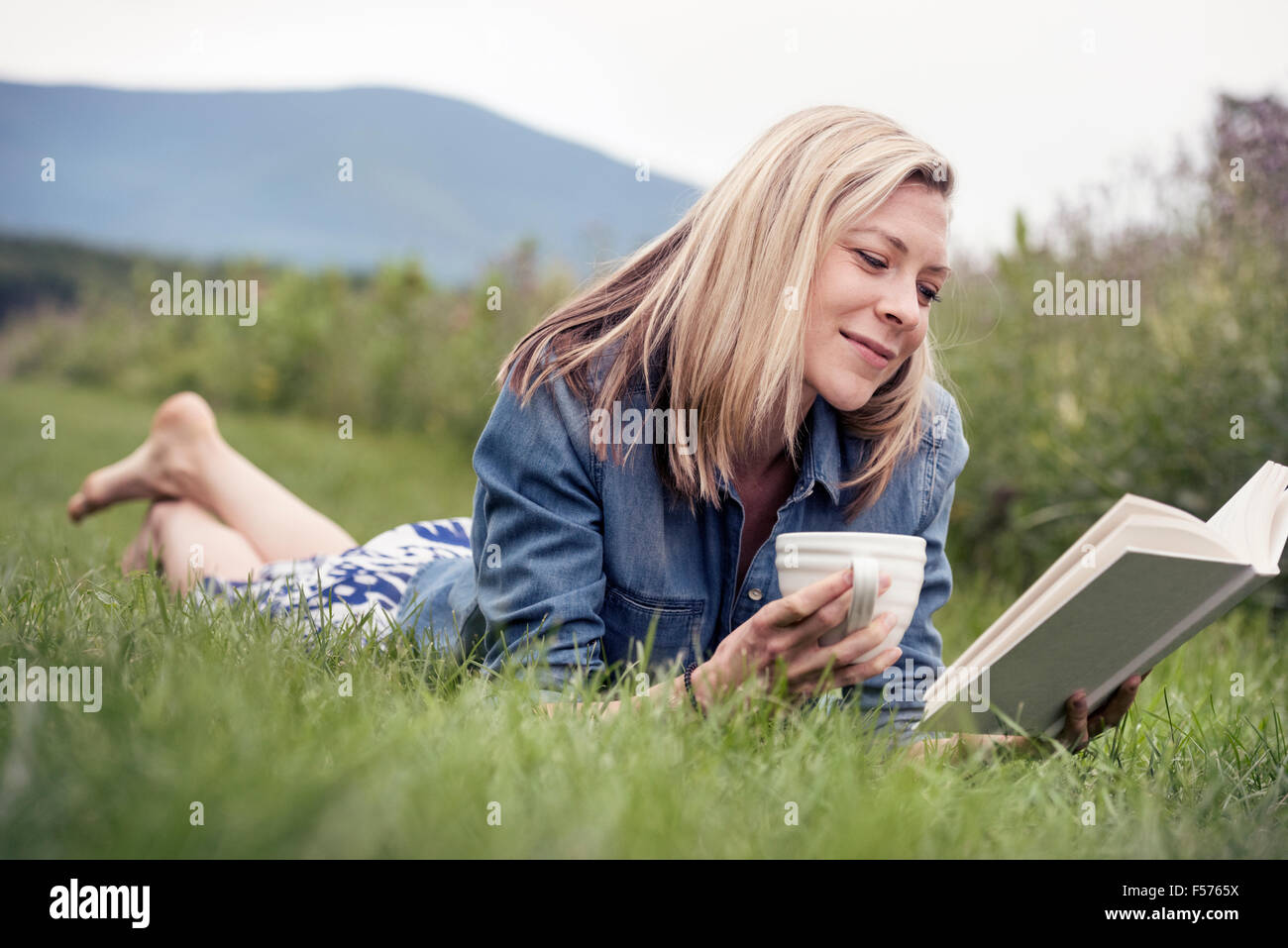 Une femme couchée sur l'herbe tenant une tasse de thé et la lecture d'un livre. Banque D'Images