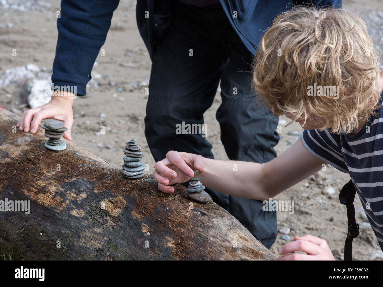 Père et fils de l'adolescence se concentrer et trouver un juste équilibre entre les cailloux, dans le style d'Andy Gray, sur un up log sur une plage Devon Banque D'Images