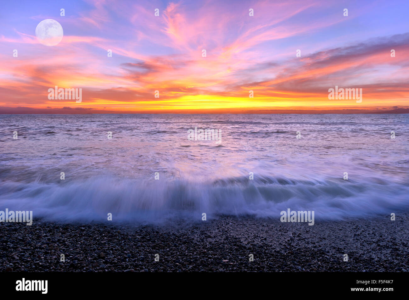 Ocean sunset lune est un ciel rempli de couleurs avec les vagues se brisant sur le rivage et la pleine lune se levant à l'arrière-plan. Banque D'Images