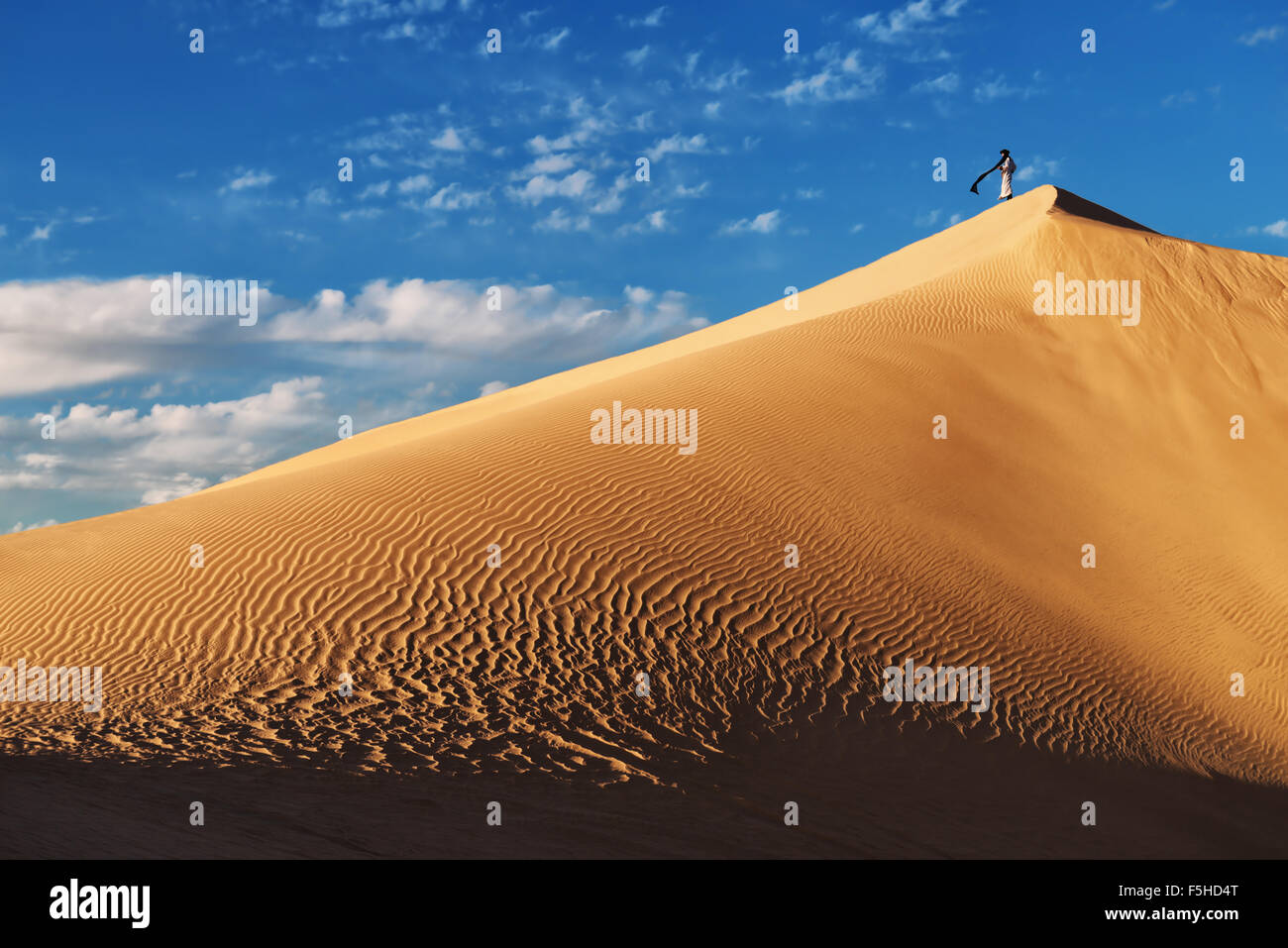 Un homme marocain traditionnel habillé se dresse sur une dune de sable contre un ciel bleu nuageux. Banque D'Images