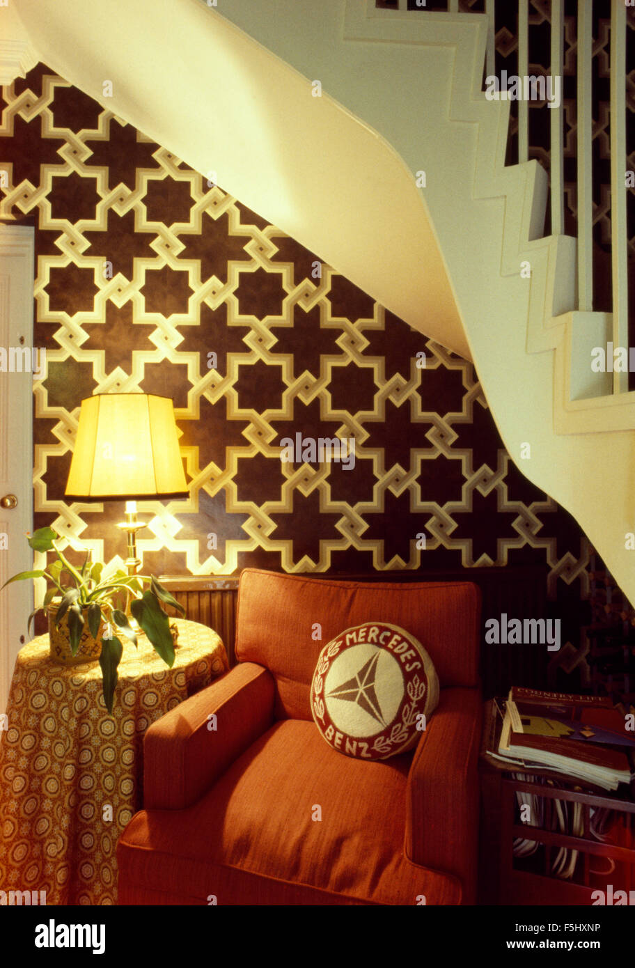 Lampe allumée et un fauteuil de couleur rouille en dessous escalier dans hall avec papier peint animé Banque D'Images
