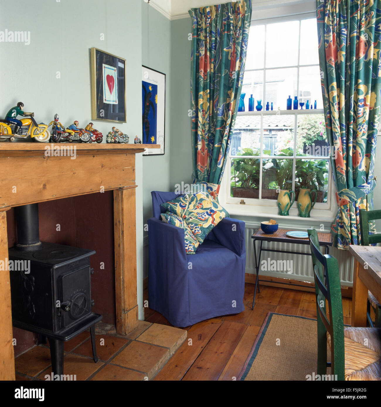 Poêle à bois dans la cheminée dans la salle à manger de style économique avec un couvercle desserré bleu sur chaise à côté de fenêtre avec des rideaux aux motifs floraux Banque D'Images